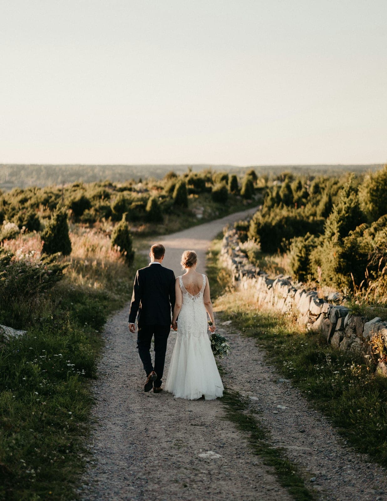 A bride and groom walking hand in hand down a gravel path through open countryside at golden hour
